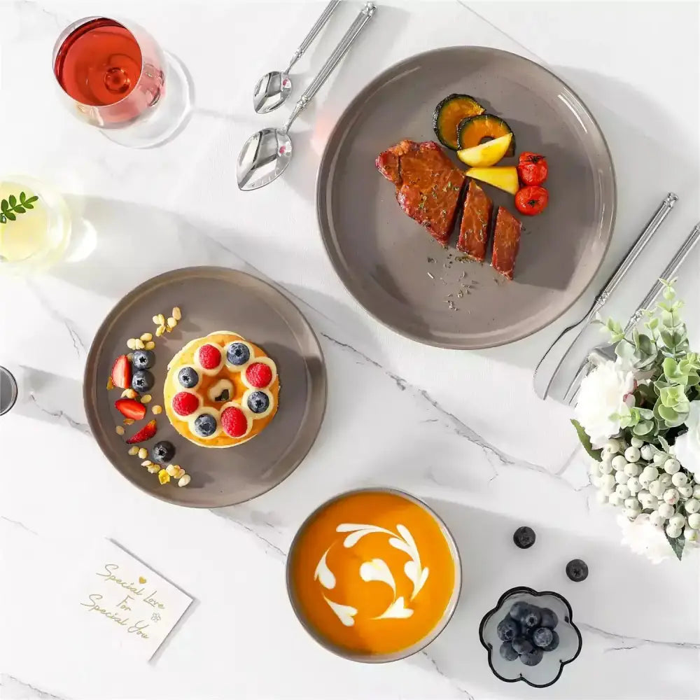 Grå keramiktallrikar och skålar med bestick, glas och blomsterarrangemang med mat